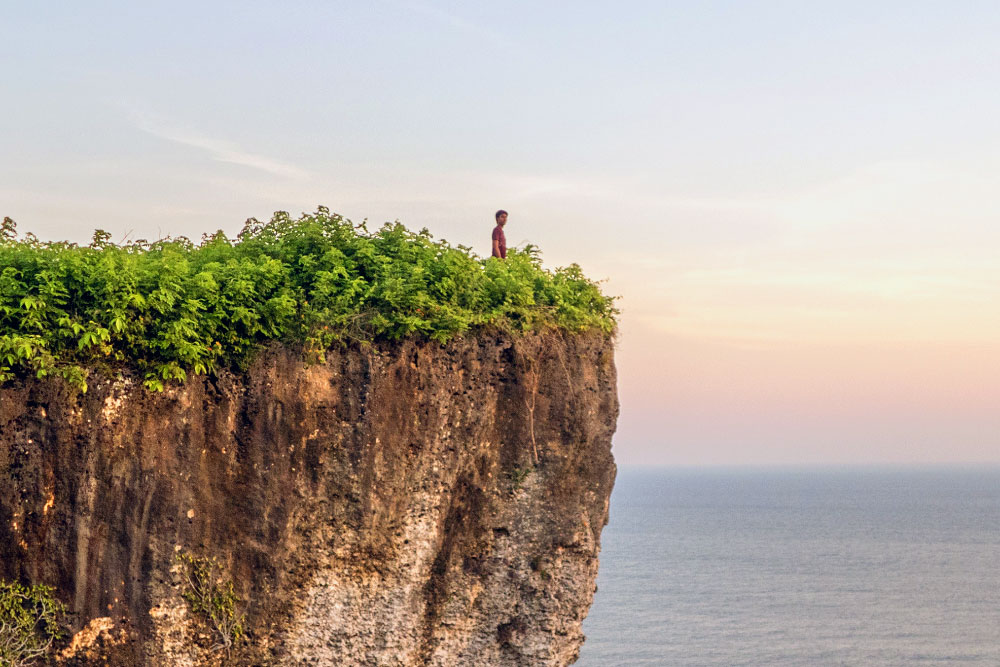 Image: A Man stands on top of a cliff, looking out at the ocean. 