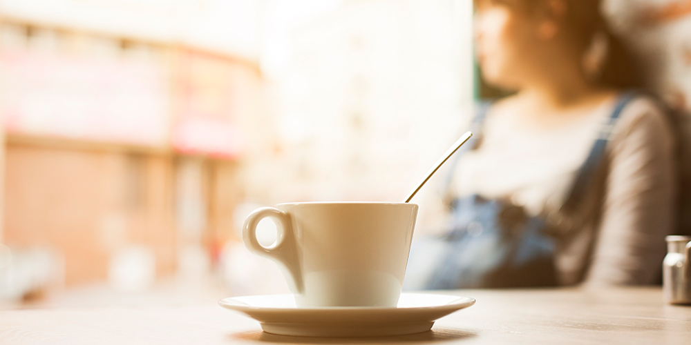 A cup of coffee in the foreground, with a girl sitting in the background. She's out of focus. 